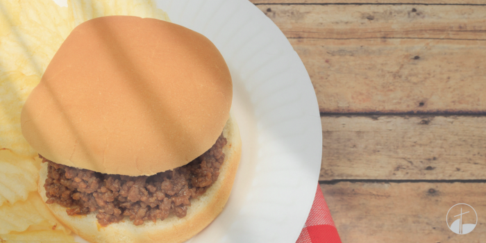 Sloppy Joe on a bun on white plate with original plain potato chips on a red and white checkered napkin.